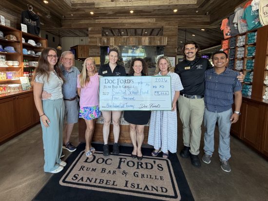 Eight people inside a shop holding a large ceremonial check from Doc Ford’s donating $4,000 to the Sanibel School Fund.