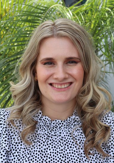 Woman with wavy light hair in a white polka‑dot blouse standing in front of green palm leaves.