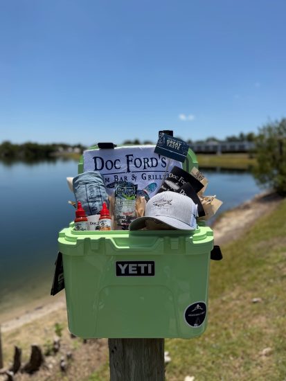 Light green YETI cooler filled with Doc Ford’s merch, snacks, and sauces, displayed outdoors by the water.