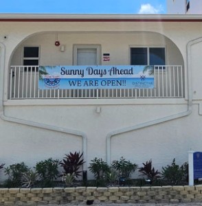 Light-colored building with balcony and a banner reading “Sunny Days Ahead — We Are Open!” above a small garden.