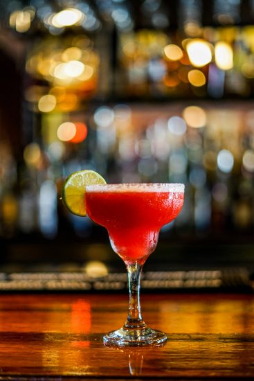 Red frozen margarita in a salted glass with a lime garnish on a bar, with colorful lights blurred behind.