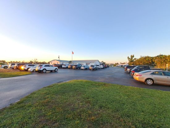 Large parking lot at sunset with rows of cars and a building labeled “Port Boat” flying an American flag.